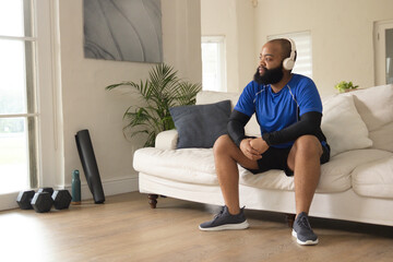 Fototapeta premium African American adult man sitting on sofa wearing headphones and blue shirt, dumbbells, copy space