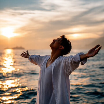 白いシャツをなびかせ夕日の海辺で両手を広げ喜びを表現する若い青年の姿