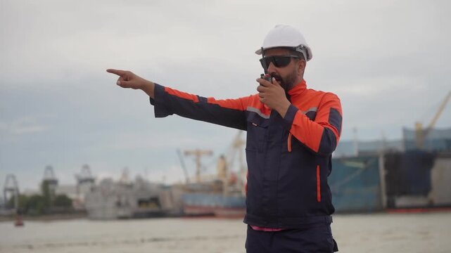 Port engineer pointing direction and speaking on walkie talkie at harbor, Marine supervisor using radio communication and pointing forward outdoors, Worker managing logistics and giving instructions