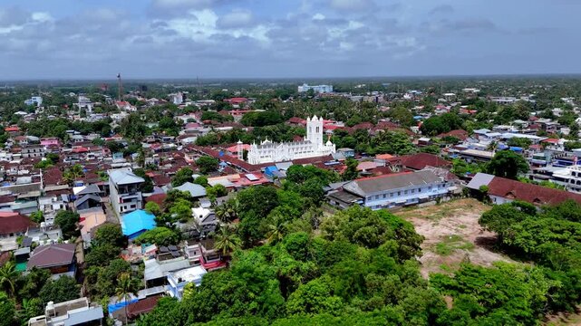 Wide aerial drone view of St. George Syro Malabar Catholic in Alappuzha, Kerala, India. Features white Gothic architecture, residential rooftops, dense green trees, and distant horizon under blue sky.