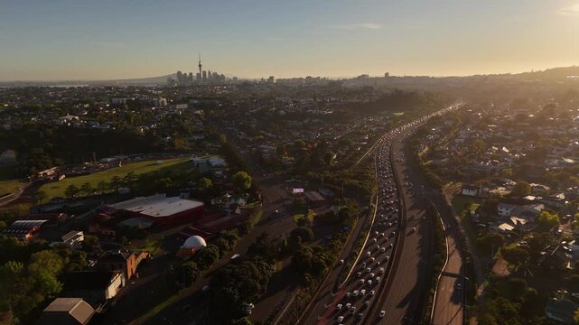 Congested motorway traffic stretches toward Auckland skyline at sunset in New Zealand, Sky Tower silhouette rises above hazy skyline as residential suburbs spread below. Aerial drone wide shot