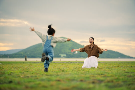 excited preschool child girl running into her mother's open arms on grass field near the river with mountain