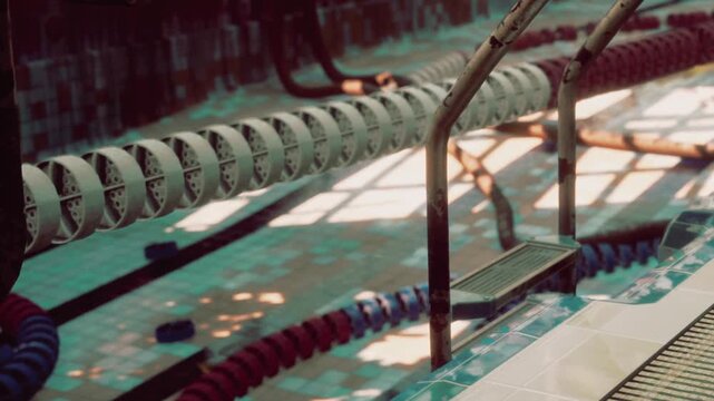 An indoor swimming pool with starting blocks. Swimmers stand near the edge, preparing for their race. Light filters through the windows above the pool.