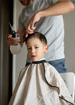 Father gives his son a haircut with scissors, comb and spray bottle at home. Young boy wears a barber cape during grooming as his dad carefully trims and styles his wet hair in a bright room.