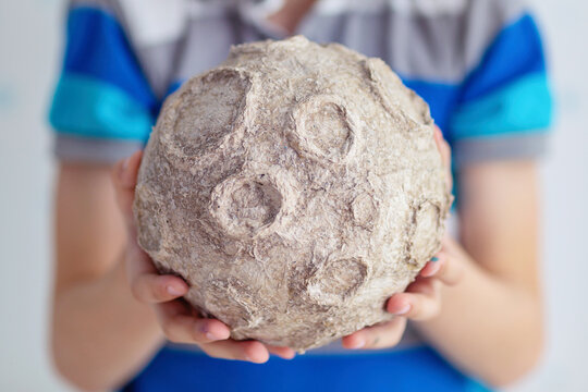 Child holds a handmade papier mache moon model with craters during a creative space education project, showing a textured lunar surface in closeup against a bright blue and white background.
