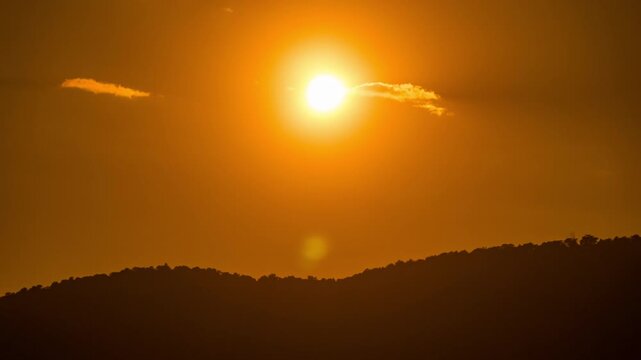 Time-lapse of the sun disappearing behind dark mountain silhouettes under a majestic sky.