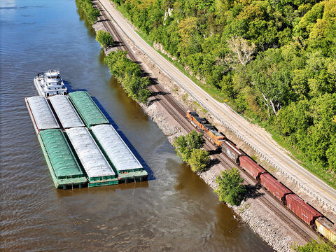 Aerial view of a tug boat with barges waiting outside the main channel next to the Mississippi shoreline while a train makes its way North