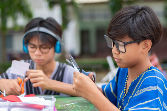 Two Asian teenage boys concentrating on cutting paper with scissors, doing arts and crafts or a school project together outdoors.