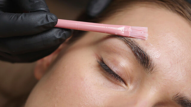 A close-up photo of a person's face during a professional eyebrow waxing or tinting treatment, with a gloved hand holding a pink brush over the eyebrow area