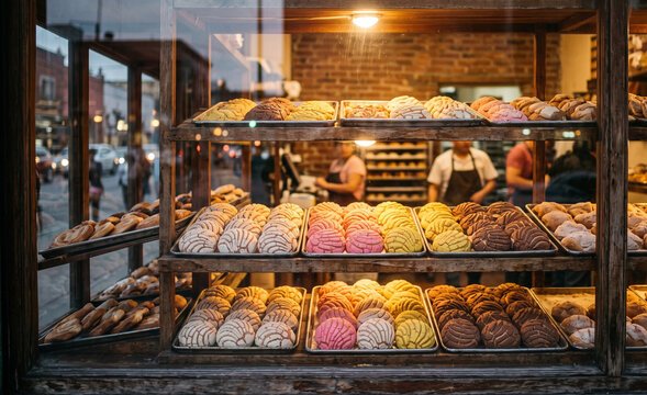 artisan bakery counter with wooden trays holding pink and chocolate concha sweet breads, Soft golden glow on sugar crusts and organized bread presentation.