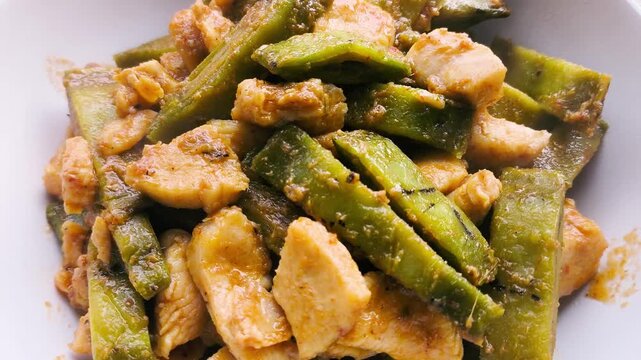 A close-up shot of a healthy homemade dish featuring stir-fried tender chicken breast chunks and sliced green pods of Oroxylum indicum (Broken Bones Tree) in a savory seasoning sauce, served in a whit