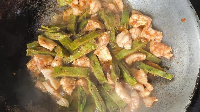  A close-up shot of cooking a unique Thai dish, stir-fried chicken breast with sliced local green fruit called Oroxylum indicum (Phayom) inside a rustic metal wok. The video captures the hot sauce bub