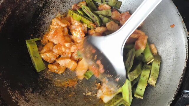  A close-up shot of cooking a unique Thai dish, stir-fried chicken breast with sliced local green fruit called Oroxylum indicum (Phayom) inside a rustic metal wok. The video captures the hot sauce bub