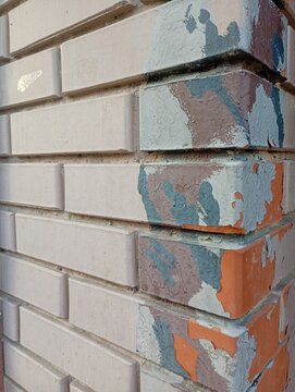 Close-up perspective view of an old brick wall corner with layers of peeling paint in light grey, dark grey, and brown tones, revealing the textured weathered red brick surface underneath.