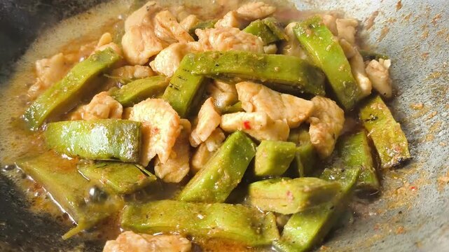  A close-up shot of cooking a unique Thai dish, stir-fried chicken breast with sliced local green fruit called Oroxylum indicum (Phayom) inside a rustic metal wok. The video captures the hot sauce bub