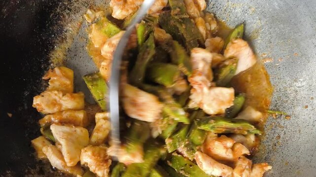  A close-up shot of cooking a unique Thai dish, stir-fried chicken breast with sliced local green fruit called Oroxylum indicum (Phayom) inside a rustic metal wok. The video captures the hot sauce bub