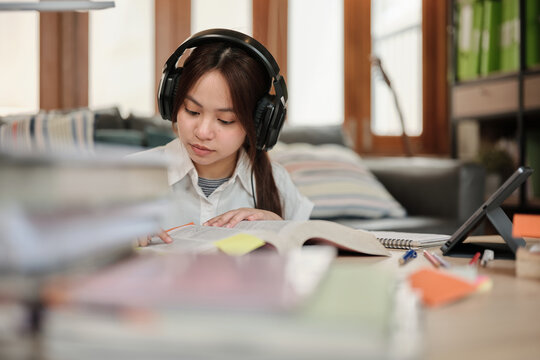 Asian teenage girl in headphones concentrates on learning at home, reading textbooks, note writing, self-tutorial exams, preparation for back to school, Homeschooling, university education lifestyle.