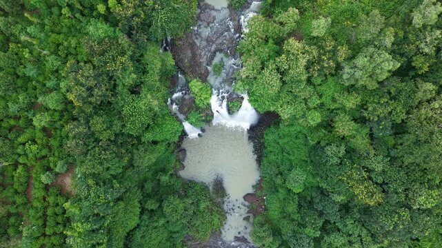 Cinematic top-down drone footage looking directly down at a spectacular three-stream waterfall as the camera gradually ascends. The widening aerial perspective progressively reveals the waterfall