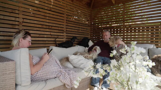 Mature married couple relaxing together on comfortable garden furniture beneath a wooden gazebo while their pet dogs gather around for affection