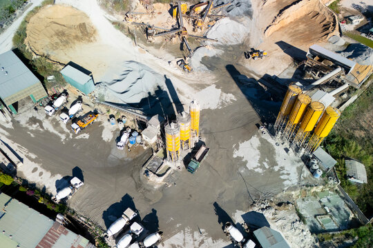 A busy concrete plant thrives with trucks and machinery moving around. Yellow silos tower over piles of gravel and sand under the bright midday sun