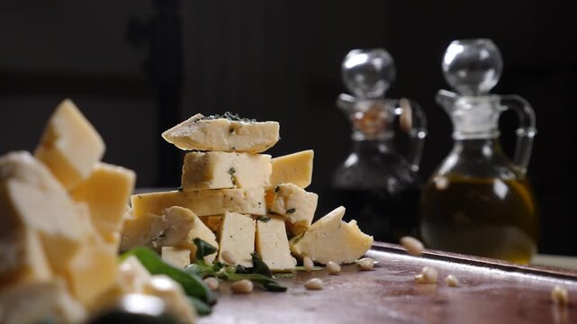 Chopped hard cheese cubes stacked pyramid on rustic wooden board. Pine nuts falling in slow motion overhead. Green herbs, glass oil vinegar bottles out of focus. Gourmet appetizer styling 4k video