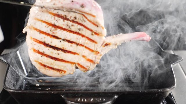 Juicy grilled pork chop lifting over cast iron skillet. Thick white smoke swirls around perfectly seared meat surface. Delicious restaurant barbecue cooking process and appetizing culinary food. 4k