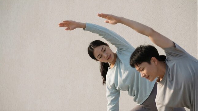 A young Asian couple practicing yoga and stretching exercises together outdoors against a minimalist neutral wall.