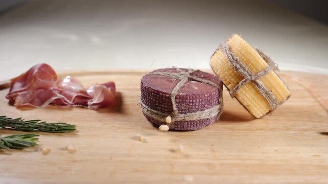 Nuts falling onto rustic wooden board displaying sliced cured meat, purple and artisan cheeses tied with twine, and fresh rosemary. Gourmet appetizer food styling and preparation. Slow motion 4k video