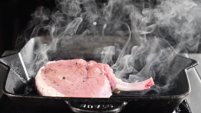 Juicy pork rib chop sizzling on hot iron skillet creating thick white smoke. Seasoned meat surface, intense cooking process. Appetizing barbecue grilling technique, gourmet dining preparation 4k