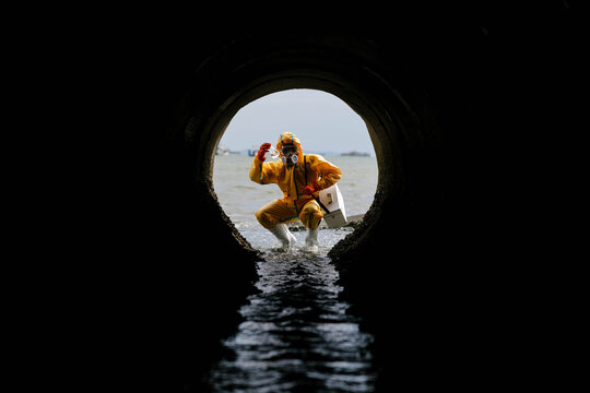 View from inside a dark drainage pipe of a waterproofed worker crouching in flowing water, inspecting the outfall toward open water, framed by the circular tunnel.