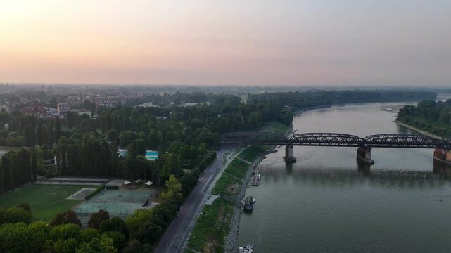 Po River and historic iron bridge connect Cremona, Lombardy, with Castelvetro Piacentino, Piacenza, Emilia Romagna, Italy, beside a wooded riverside promenade and urban waterfront, drone forward flyov