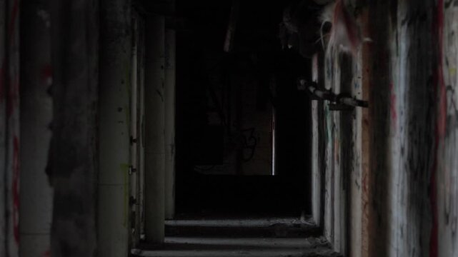 Light enters through broken windows in a dark abandoned factory corridor, revealing graffiti, decay and a spooky, unsettling industrial interior, static tripod shot