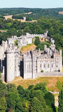Arundel Castle drone orbit reveals medieval stone fortress, towers and walls above wooded parkland in West Sussex, England, on a clear summer day with rolling hills beyond. Vertical format.