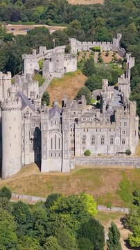 Arundel Castle aerial flyover during summer heatwave, revealing drought-browned lawns, historic stone walls and towers, and dense tree canopy surrounding the landmark in West Sussex, England. Vertical
