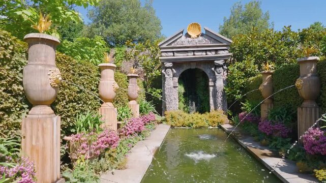 Arundel Castle gardens: rightward pan along a narrow water rill with fountain jets toward a classical stone grotto pavilion, flanked by urns, clipped hedges and pink flowering borders.