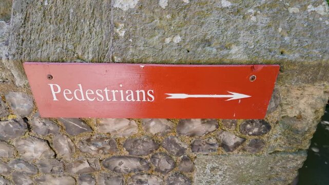 Pedestrians sign with right arrow on red plaque fixed to rough stone wall; camera pans right to show walkway, outdoor wayfinding and public access direction in daylight.