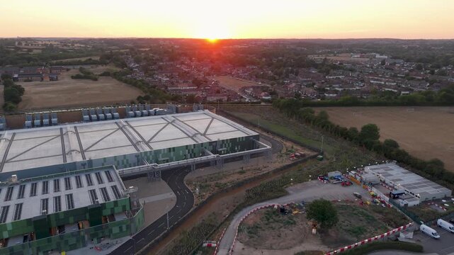 Google data centre Waltham Cross at sunset as drone reverses and pans left over the campus, revealing rooftops, service roads and surrounding fields with the sun on the horizon.