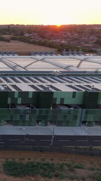 Google data centre, Waltham Cross, England. Right pan aerial view of large campus building at sunset, golden hour light with sun low on the horizon behind. Vertical format.