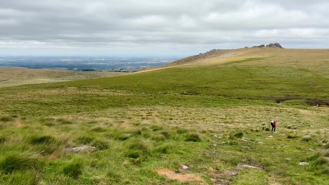 Moorland ridge above Two Bridges in Dartmoor National Park England with rolling grassland distant hills cloudy sky and a lone hiker crossing the landscape