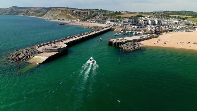 Aerial drone View of West Bay Harbour in Dorset England showing boat entering between stone piers with sandy beach, coastal cliff and marina along Jurassic Coast