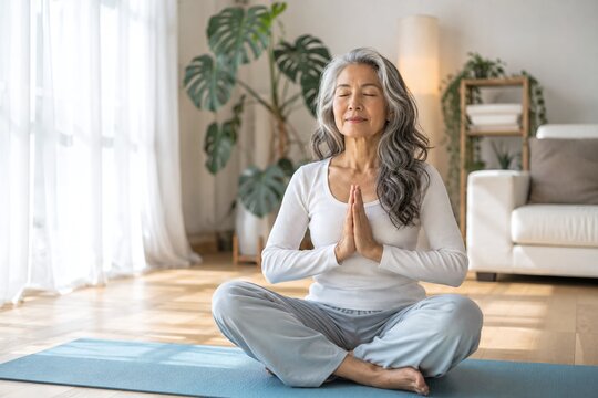 Peaceful senior Asian woman with long grey hair practicing meditation in namaste pose at home, mature female doing yoga on mat in bright living room, wellness and healthy aging concept