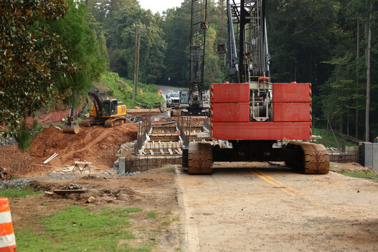 Heavy crawler crane and machinery operating at an infrastructure construction site surrounded by trees.