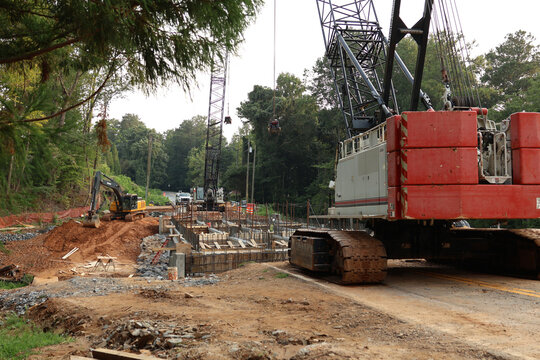 Heavy crawler crane and machinery operating at an infrastructure construction site surrounded by trees.