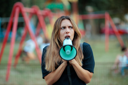 Angry Young Mother Shouting Through Megaphone At Children Playground. An upset mom uses a loud megaphone to call kids at park