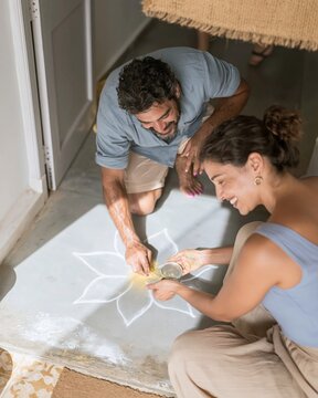 Bengali man and woman creating a colorful rangoli pattern on a cement threshold, showing quiet concentration for festive preparation and craft