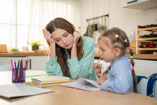 Tired mother sitting at kitchen table looking stressed while her young daughter doing homework with color pencils. Exhausted mother or tutor looking bored while helping little girl with home schooling