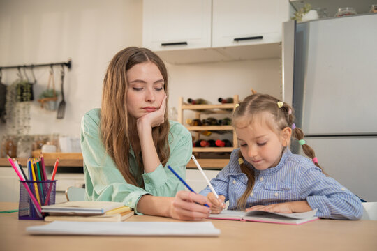 Mother guiding her little daughter's hand while writing homework at home kitchen table. Young mother helping daughter write in notebook during home study, showing care and patience despite being tired