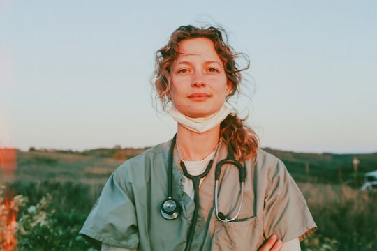 Nurse in green scrubs standing outdoors at sunrise with a weary steadfast gaze, featuring a face mask pulled down and a stethoscope for healthcare recruitment