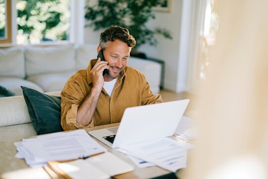 Mature businessman working at a kitchen table while talking on a phone, natural home office scene with a laptop and papers for small business marketing