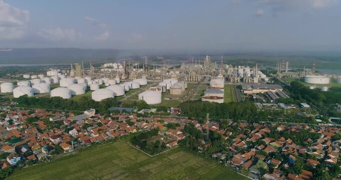 Aerial view of the oil refinery complex with storage tanks and industrial infrastructure adjacent to residential areas and green fields in Cilacap Regency, Central Java, Indonesia.